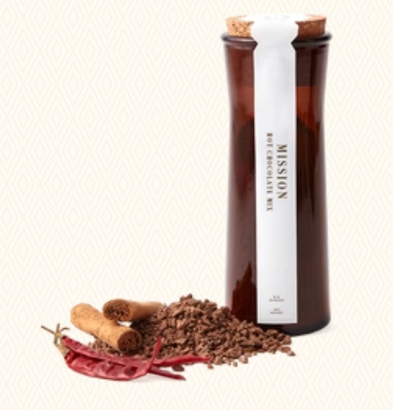 A Mission Hot Chocolate Mix jar with a white label and cork stopper, displayed next to loose chocolate chips, cinnamon sticks, and red chili peppers on a light beige patterned background.