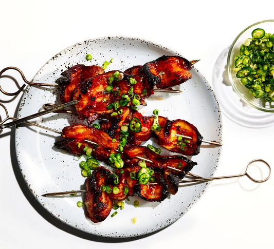Grilled bacon kebabs on skewers, garnished with sliced green onions, on a white plate