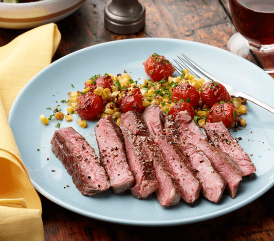 Sliced steak with roasted tomatoes and corn on a blue plate, served with a glass of red wine.