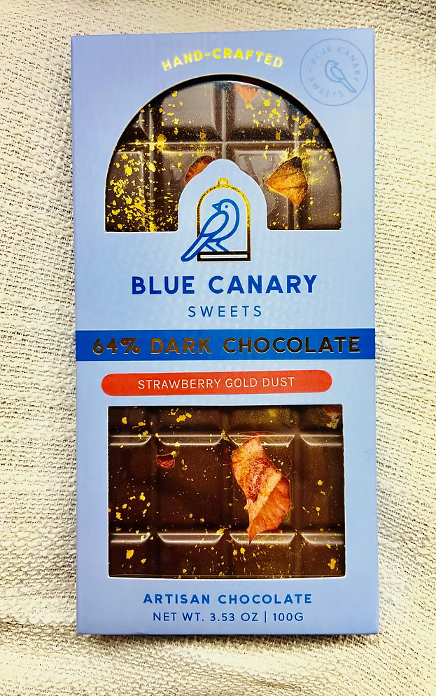 Dark chocolate bar with strawberries and gold dust, wrapped in blue cardboard on a cream burlap background.