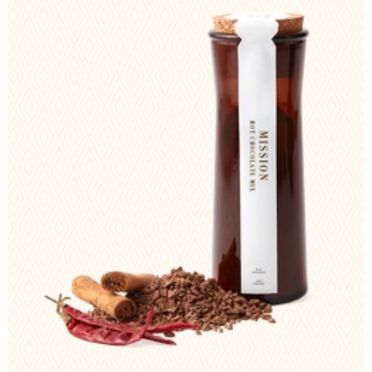 A Mission Hot Chocolate Mix jar with a white label and cork stopper, displayed next to loose chocolate chips, cinnamon sticks, and red chili peppers on a light beige patterned background.
