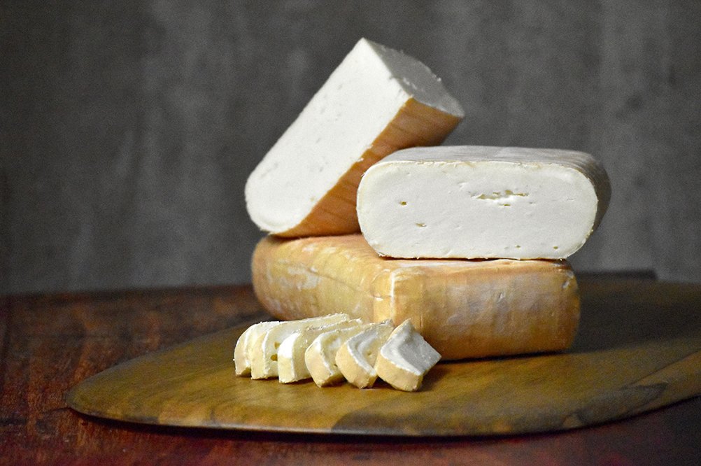 Several wedges and slices of soft white cheese (Jasper Hill's Eligo) stacked on a round wooden board against a dark textured studio background.